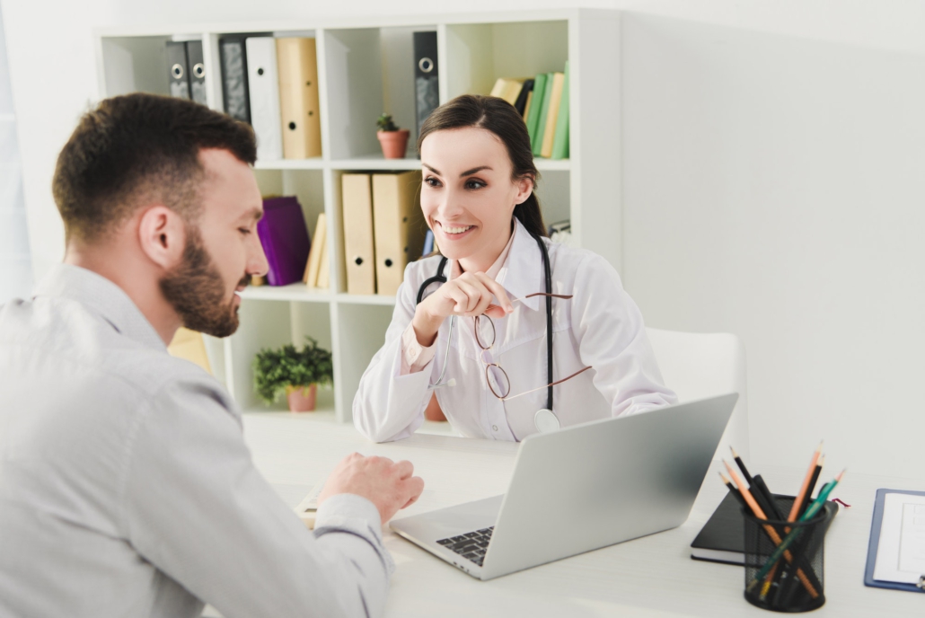 smiling doctor and man discussing health insurance and looking on laptop screen in clinic